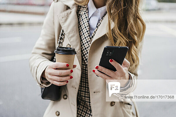 Mittlere erwachsene Frau  die ein Mobiltelefon benutzt und einen Einweg-Kaffeebecher auf der Straße hält