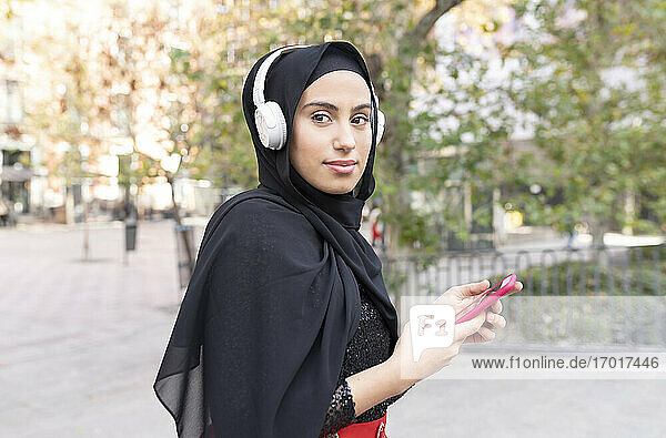 Porträt einer jungen schönen Frau mit Hijab und Kopfhörern  die ein Smartphone benutzt