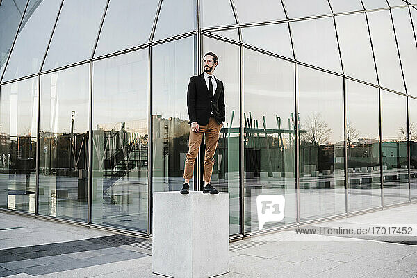 Geschäftsmann schaut weg  während er auf einer Bank vor einem modernen Gebäude steht