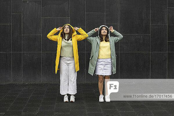 Junge Frauen in Regenmänteln schauen nach oben  während sie an einer schwarzen Wand stehen