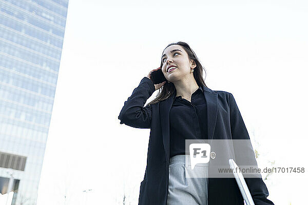 Young businesswoman with laptop looking away while talking on mobile phone standing against sky