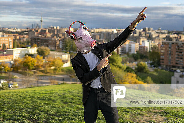 Männlicher Unternehmer mit Schweinemaske und drahtlosen Kopfhörern posiert auf einem Hügel vor der Stadt