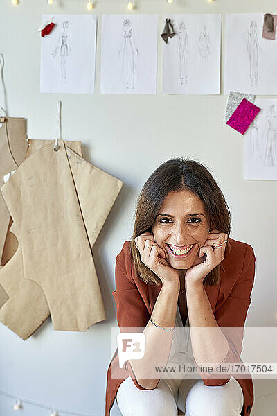 Fröhliche Modedesignerin sitzt mit den Händen am Kinn vor ihren Entwürfen und Mustern an einer weißen Wand im Atelier
