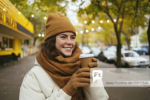 Glückliche Frau mit Strickmütze träumt mit Einweg-Kaffeebecher auf der Straße im Winter