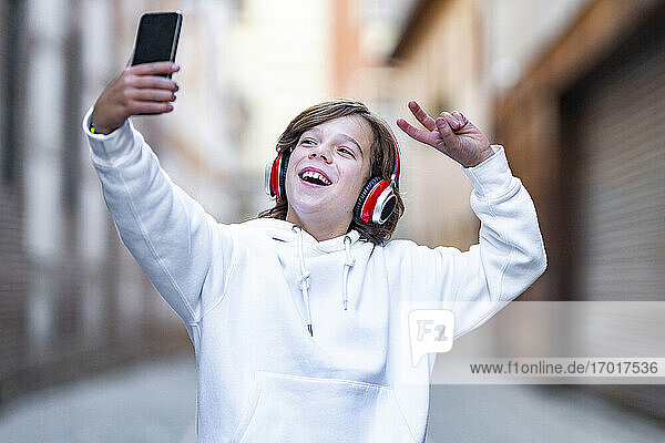 Lächelnder Junge mit Kopfhörern  der eine Friedensgeste zeigt  während er ein Selfie mit seinem Mobiltelefon im Freien macht