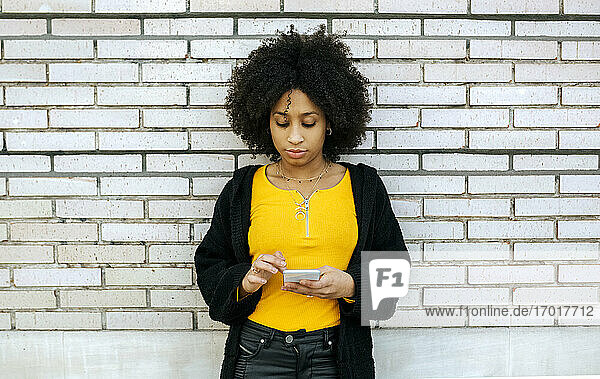 Schöne Frau mit Afro-Haar  die ein Smartphone benutzt  während sie an einer Mauer steht