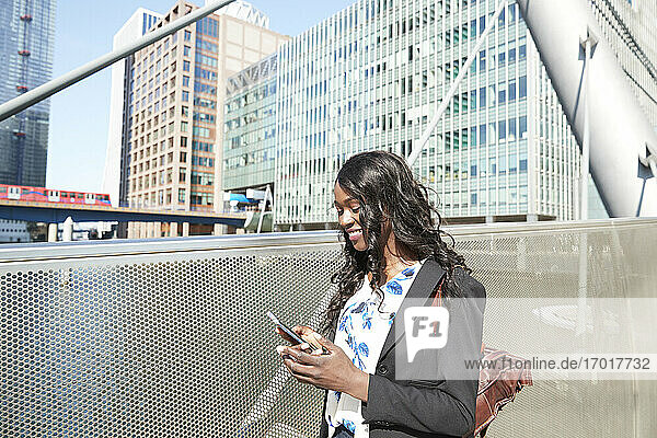 Lächelnde Geschäftsfrau auf einer Brücke  die an einem sonnigen Tag ein Mobiltelefon benutzt