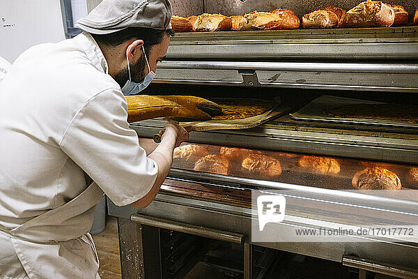 Männlicher Bäcker mit Pizzaschieber bei der Kontrolle von Brot im Ofen in einer Bäckerei während COVID-19