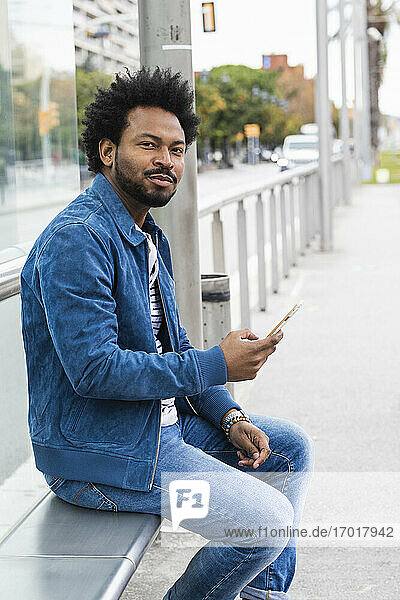 Selbstbewusster Mann mit Afro-Haar  der ein Mobiltelefon benutzt  während er auf einem Sitz an einer Bushaltestelle sitzt