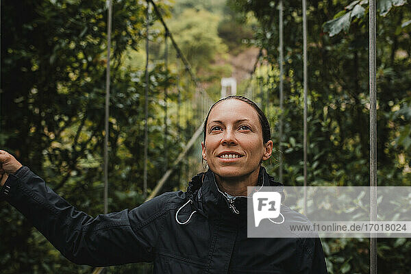 Nachdenkliche Frau im Regenmantel auf einer Hängebrücke im Wald stehend