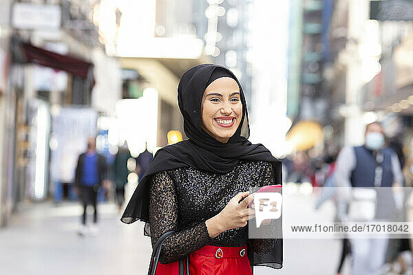 Porträt der jungen schönen Frau mit schwarzem Hijab lächelnd auf dem Bürgersteig mit Smartphone in den Händen