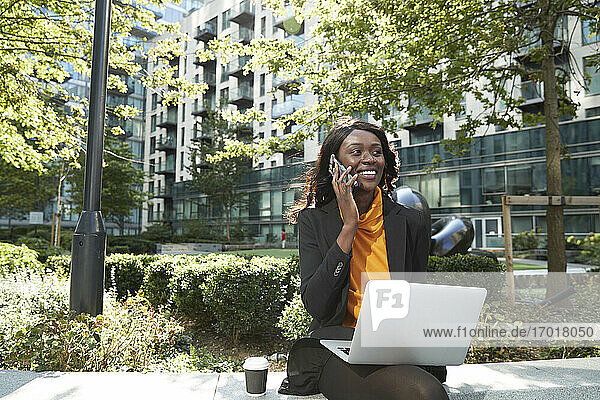 Smiling businesswoman with laptop talking on phone at office park