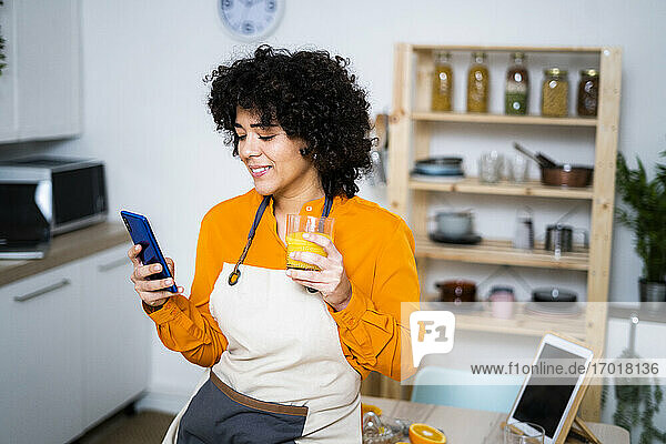Junge Frau mit Orangensaft und Mobiltelefon  während sie zu Hause auf dem Tisch sitzt