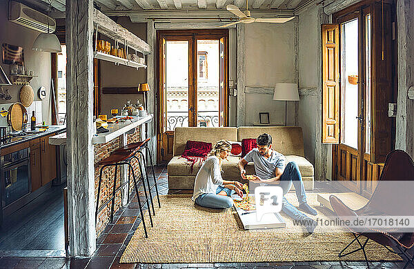 Junges Paar sitzt auf dem Boden des Wohnzimmers und isst Pizza aus einem Karton