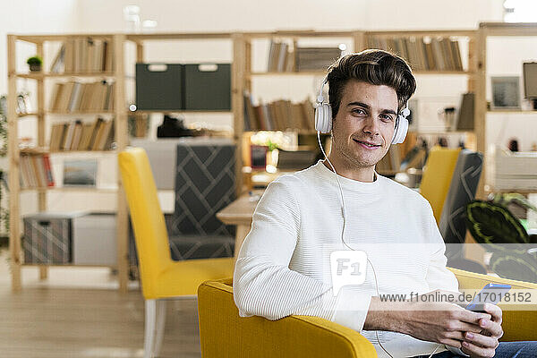 Smiling young man listening music through headphones while holding mobile phone in new home