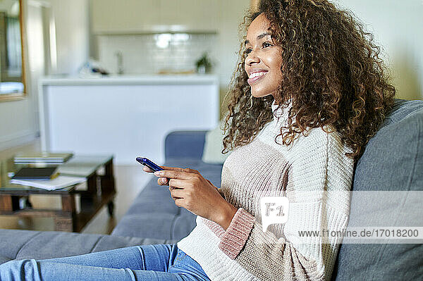 Lächelnde junge Frau mit Mobiltelefon träumt im Wohnzimmer zu Hause