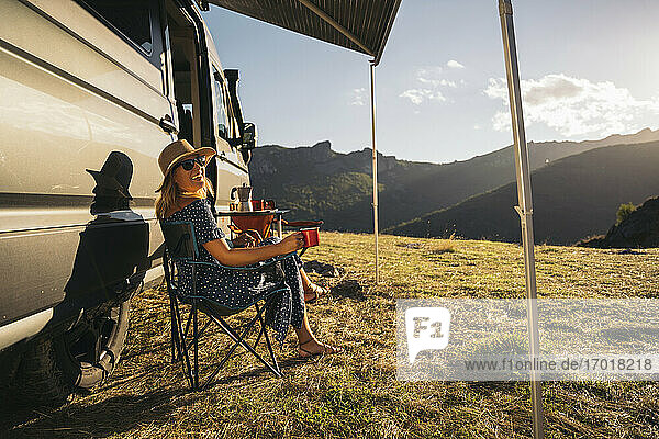 Glückliche Frau mit Sonnenbrille und Kaffeetasse  die lachend am Wohnmobil sitzt  während sie Urlaub macht