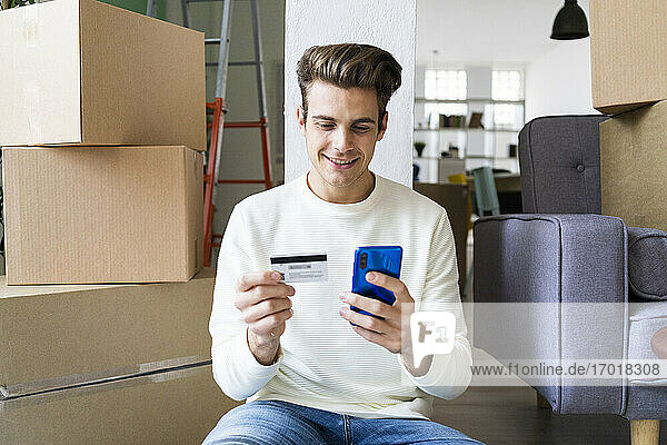 Lächelnder junger Mann  der beim Einzug in ein neues Haus mit dem Mobiltelefon und der Kreditkarte bezahlt