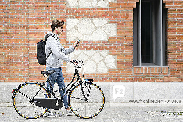 Smiling man on video call wheeling bicycle by building