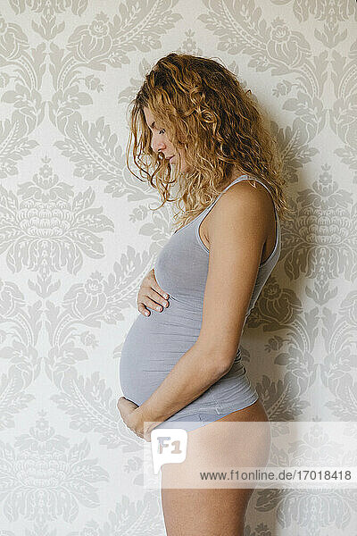 Pregnant woman caring by touching abdomen while standing against wall at home