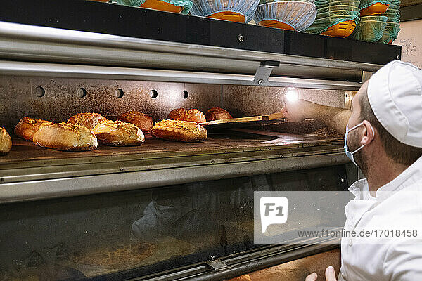 Männlicher Koch nimmt gebackenes Brot mit Pizzaschieber aus dem Ofen in einer Bäckerei