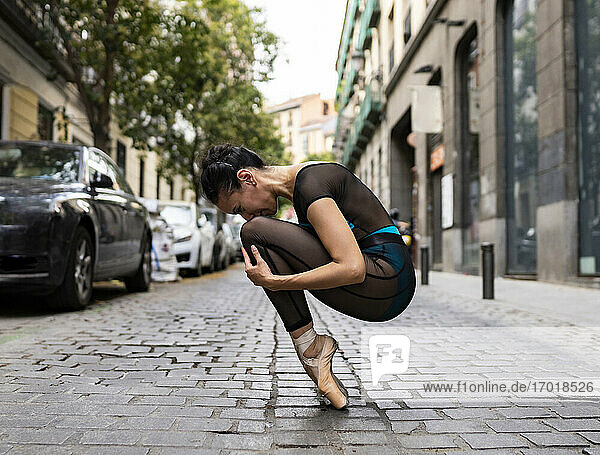 Ballet dancer wearing leotard crouching on footpath in city