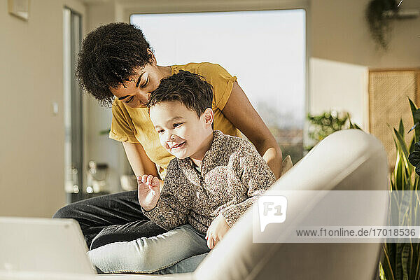 Smiling boy waving hand to video call on digital tablet while sitting by woman at home