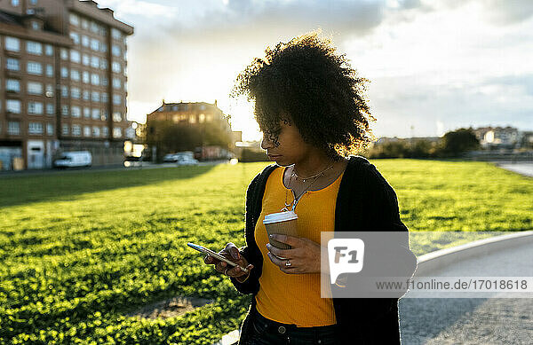 Junge Frau hält Kaffee und benutzt ein Mobiltelefon  während sie auf einem Fußweg gegen den Himmel läuft