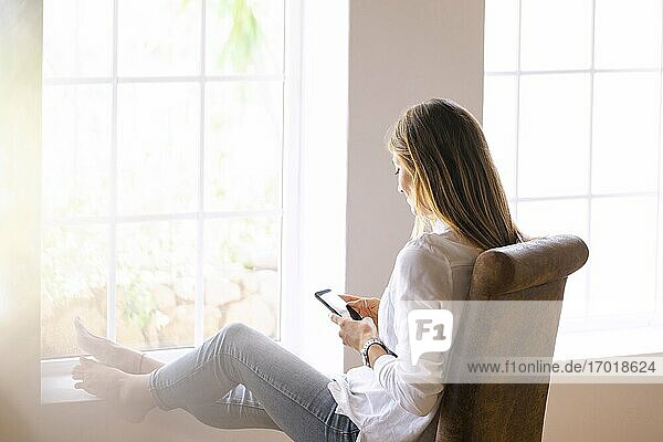 Junge Frau  die ihr Smartphone benutzt  während sie auf einem Stuhl sitzt und sich zu Hause am Fenster entspannt