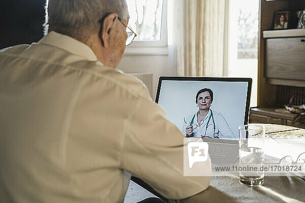 Eine Ärztin berät einen älteren Mann am Laptop per Videoanruf im Wohnzimmer