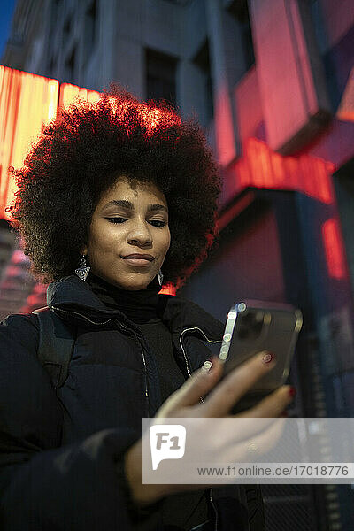 Afrojunge Frau in warmer Kleidung  die nachts in der Stadt ihr Smartphone benutzt