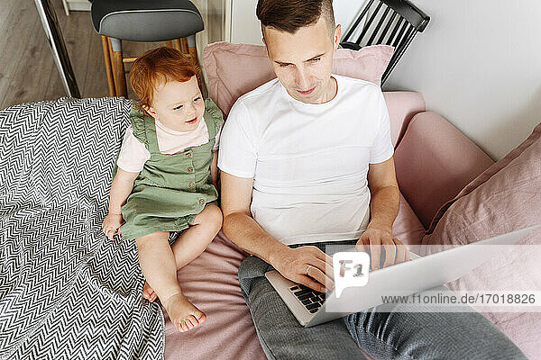 Man with baby daughter working on laptop on sofa