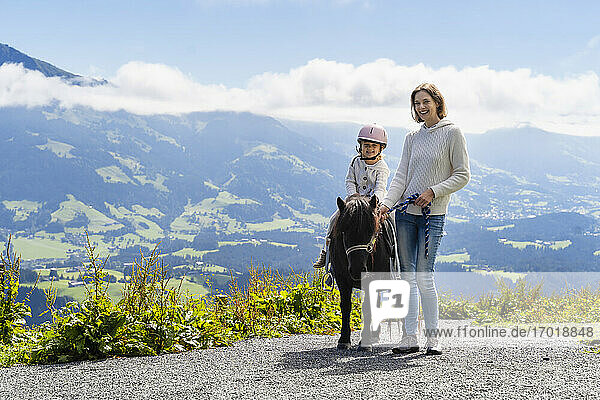 Mutter steht neben ihrer kleinen Tochter  die auf einem Pony gegen ein Bergtal reitet