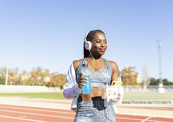 Lächelnde Sportlerin  die wegschaut  während sie Musik hört und einen Energydrink und ein Smartphone gegen den klaren Himmel hält