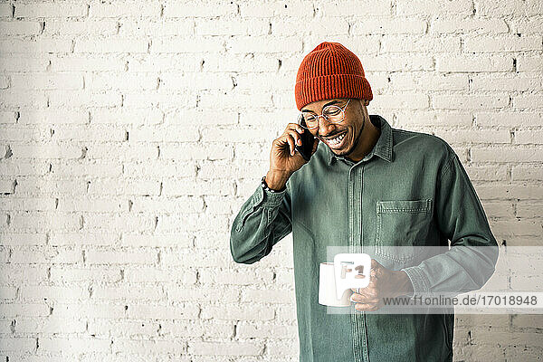 Lächelnder Mann mit Strickmütze telefoniert und hält eine Kaffeetasse an eine weiße Backsteinwand