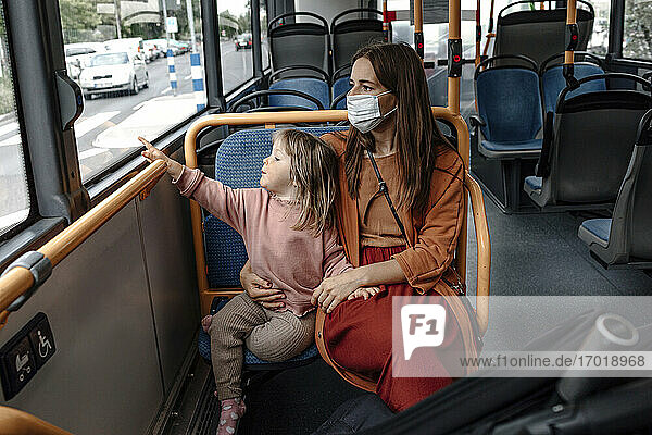 Mother with cute daughter pointing at window while traveling by bus during COVID-19