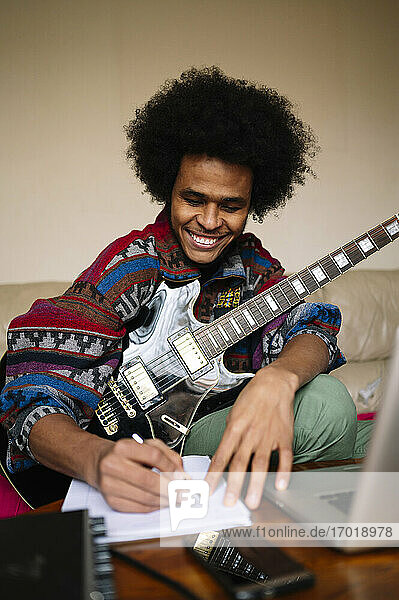 Glücklicher männlicher Gitarrist mit Gitarre schreibt Noten in ein Buch auf dem Tisch zu Hause
