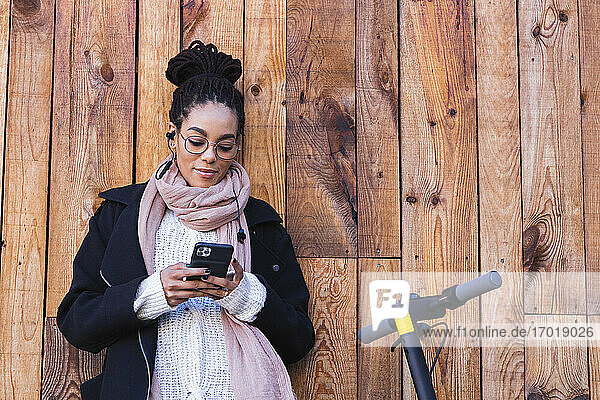 Junge weibliche Millennials benutzen ihr Smartphone  während sie sich mit dem Elektroroller gegen eine Holzwand lehnen