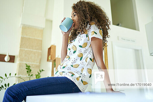 Afro junge Frau mit lockigem Haar trinkt Kaffee  während sie zu Hause auf dem Küchentisch sitzt