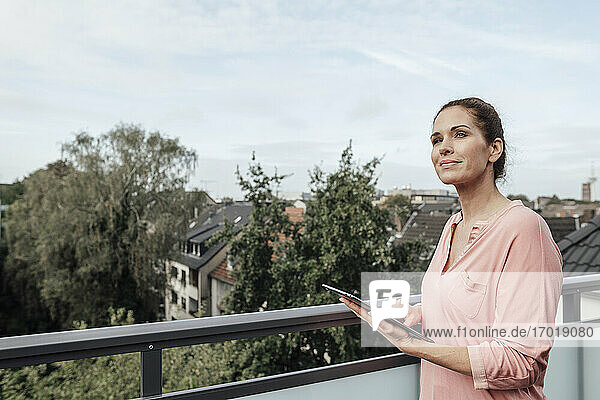 Lächelnde reife Frau hält digitales Tablet  während sie auf dem Balkon träumt