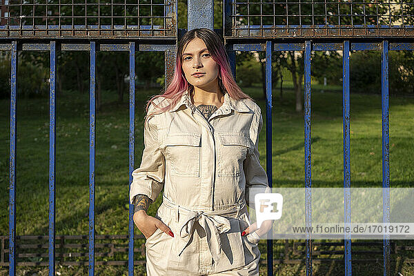 Stilvolle junge Frau mit Händen in den Taschen am Zaun an einem sonnigen Tag