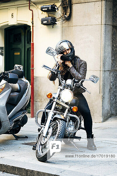 Motorradfahrerin beim Befestigen des Sturzhelms  während sie auf dem Motorrad am Bürgersteig sitzt