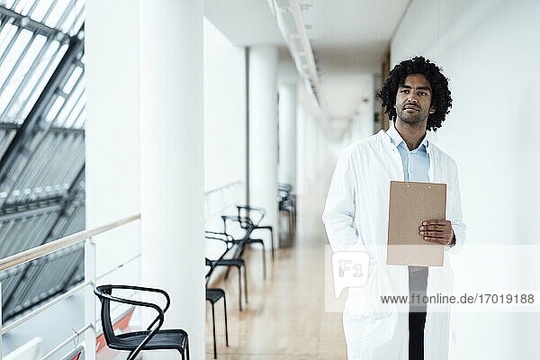 Nachdenklicher junger Mann im Gesundheitswesen  der ein Klemmbrett hält  während er in einem Krankenhaus auf dem Flur steht