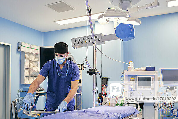 Männlicher Assistent mit Gesichtsmaske beim Einstellen des Operationstisches im Operationssaal eines Krankenhauses