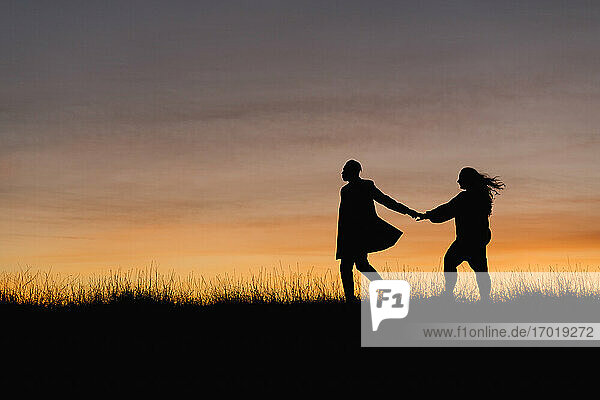 In silhouette of man holding hand of woman while walking against sky during sunset