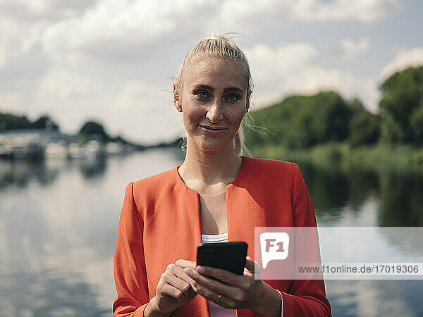 Confident businesswoman with smart phone against lake on sunny day