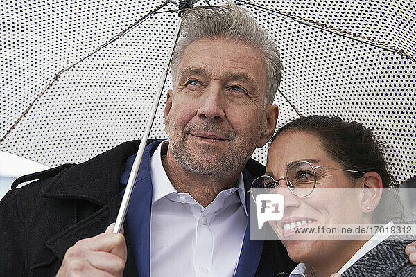 Lächelnde Geschäftsfrau mit älterem Geschäftsmann unter Regenschirm in der Regenzeit stehend
