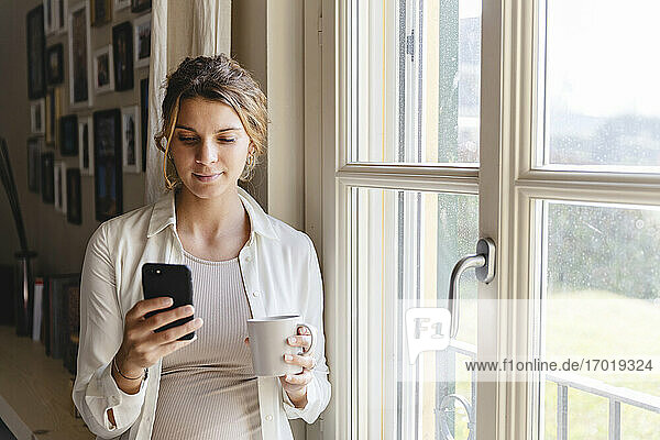 Junge schwangere Frau benutzt ihr Smartphone am Fenster zu Hause