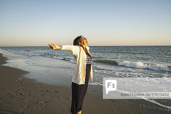 Unbekümmerte junge Frau am Strand stehend mit ausgestreckten Armen bei Sonnenuntergang