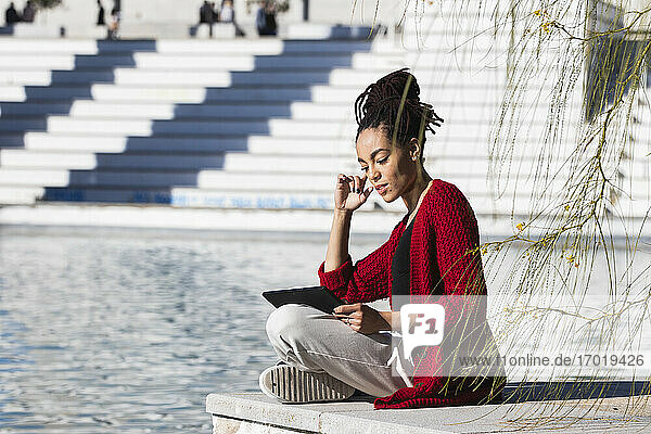 Junge Frau sitzt im Schneidersitz und benutzt ein digitales Tablet  während sie an einem sonnigen Tag am Rande einer Promenade über einem Fluss steht